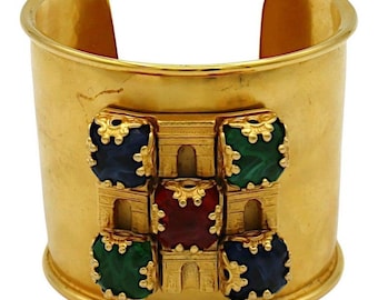 Op de afbeelding: Gouden armband met een decoratief ontwerp met een vierkant van vijf gekleurde stenen. De stenen zijn rood, blauw en groen.