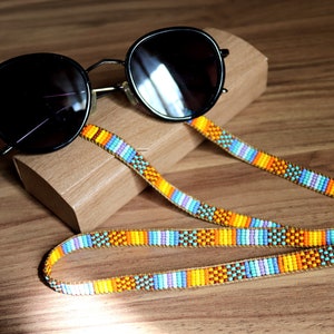 May include: A colorful beaded glasses chain with orange, yellow, blue, and purple beads. The chain is lying on a wooden surface with a pair of black sunglasses and a wooden case.
