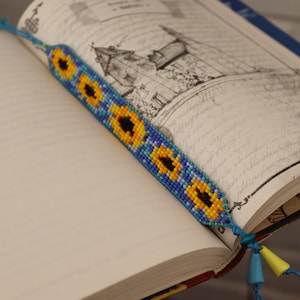 Op de afbeelding: Een blauwe en gele kralenboeklegger met een zonnebloemontwerp. De boeklegger ligt op een boek met een tekening van een gebouw.