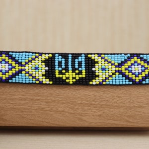 May include: A blue, yellow, white and black beaded bracelet with a trident design.