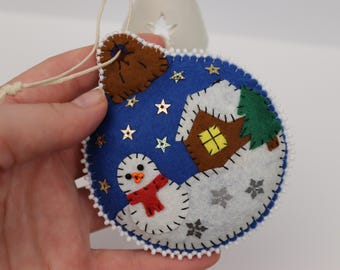 May include: A blue felt Christmas ornament with a snowman, house, and tree design. The ornament has a white beaded edge, gold star accents, and a brown top. A red scarf is around the snowman's neck.