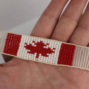 May include: A beaded bracelet featuring the Canadian flag design. The bracelet has a white background with a red maple leaf in the center. Red stripes border the maple leaf, and the ends have a gradient of white and gray beads.
