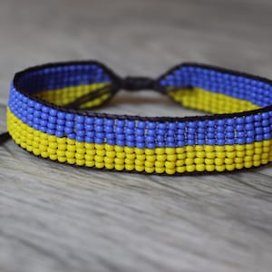 Può includere: Un braccialetto di perline con perline gialle e blu, che rappresentano la bandiera ucraina. Il braccialetto è legato con un cordoncino nero.
