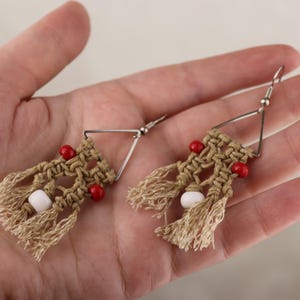 Puede incluir: Pendientes de macramé hechos a mano con un diseño geométrico. Los pendientes presentan un patrón beige tejido, acentuado con cuentas rojas y blancas, y ganchos de metal plateado. La borla añade un toque bohemio.