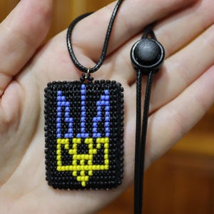 May include: A black beaded pendant necklace featuring the Ukrainian coat of arms in blue and yellow. The pendant is square-shaped and hangs from a black cord with an adjustable clasp. The design is a stylized trident.