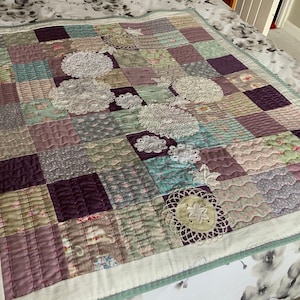 May include: A patchwork quilt featuring a mix of floral and patterned fabrics in shades of purple, green, and white. The quilt is adorned with white lace doilies.