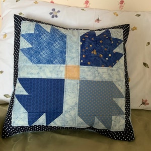 May include: A blue and white patchwork cushion with a black and white polka dot border. The cushion is made of different fabrics with various patterns, including a blue plaid, a blue with gold dots, and a blue with white dots.