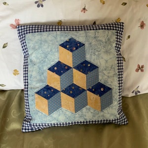 May include: A blue and beige patterned cushion with a geometric design of cubes. The cushion has a blue and white gingham border.