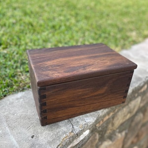 May include: A dark brown wooden box with a hinged lid. The box is made of solid wood and has a natural finish. The box is sitting on a stone surface.