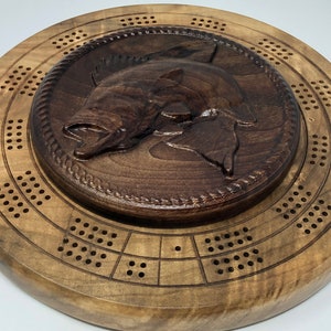 Op de afbeelding: Een ronde houten cribbagebord met een uitgesneden visontwerp in het midden. De vis is donkerbruin en het bord heeft meerdere rijen gaten voor de pinnen. De buitenste ring is lichtbruin en het bord is van hout.