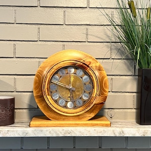 May include: A round wooden clock with a light brown finish. The clock face has a gold center and silver-colored hour markers. The clock sits on a wooden base. Decorative candles and a black vase with greenery are on either side.