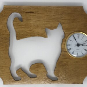 May include: Wooden wall clock featuring a white cat silhouette against a gold-toned background. The clock has a round face with Roman numerals and gold-colored trim. The cat's tail is curved.