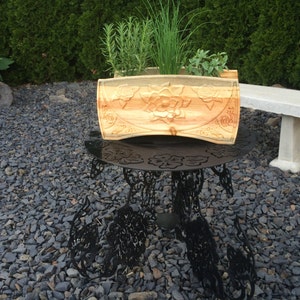 May include: A wooden planter box with carved floral designs, filled with herbs. The light-colored wood contrasts with the green plants. The planter sits on a black metal table with decorative cutouts, outdoors.