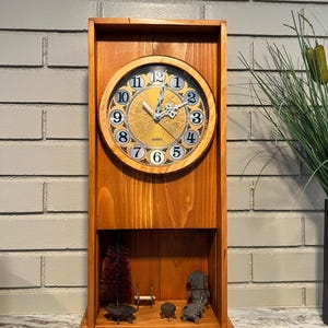 May include: A wooden clock with a round face featuring Roman numerals and decorative gold accents. The clock is housed in a rectangular wooden frame with a small display area at the bottom, containing miniature furniture and a small tree.
