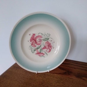May include: A white ceramic plate with a light teal rim and a floral design. The center of the plate features pink and red lilies with green leaves. The plate is displayed on a wooden surface.