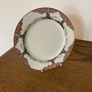 May include: A white ceramic plate with a black and orange tree design around the rim. The trees have red circles on the branches, resembling apples.