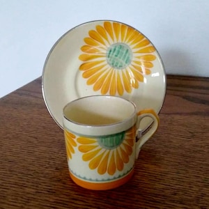 Puede incluir: Un juego de taza y platillo vintage con un diseño floral amarillo y verde. Tanto la taza como el platillo están decorados con un patrón de margarita estilizado.
