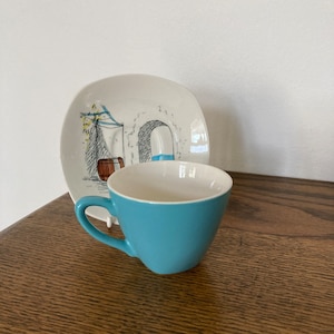 Könnte beinhalten: Eine blaue Teetasse mit weißem Innenraum und einer weißen Untertasse mit einer Schwarzweißillustration eines Gebäudes mit einem Torbogen.