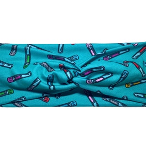 May include: A teal headband with a repeating pattern of colourful test tubes. The test tubes are in various colours, including red, green, purple, and yellow, with grey and white accents. The headband is twisted at the top.