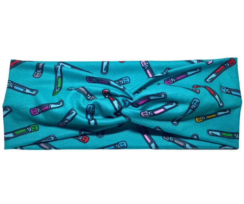 May include: A teal headband with a repeating pattern of colourful test tubes. The test tubes are in various colours, including red, green, purple, and yellow, with grey and white accents. The headband is twisted at the top.