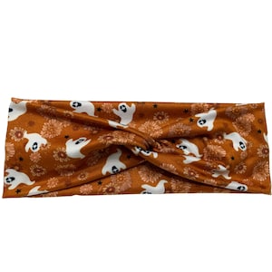 May include: A burnt orange headband with a repeating pattern of white ghosts, flowers, and black stars. The headband is twisted at the top. The fabric has a soft, matte finish.