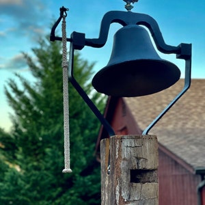 May include: A black metal bell with a rooster weathervane on top, mounted on a weathered wooden post. A braided rope hangs from the bell. The bell is set against a backdrop of green trees and a red building.