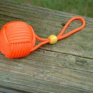 May include: A bright orange paracord keychain with a yellow bead. The keychain is made of a thick, braided cord and is tied into a knot at the end.