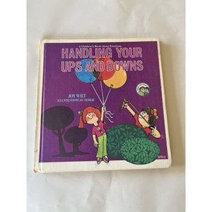 May include: A purple book cover with the title "Handling Your Ups and Downs" in white text. The book is about emotions and features illustrations of two children, one holding balloons and the other holding a slingshot.