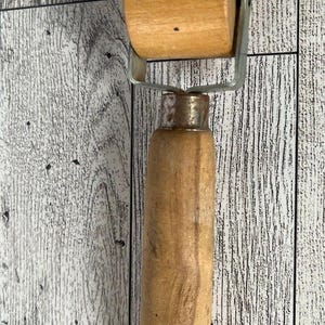 May include: A wooden seam roller with a cylindrical roller and a handle. The roller is attached to a metal frame. The handle is made of wood and has a rounded end. The tool is used for pressing seams in sewing and crafting.