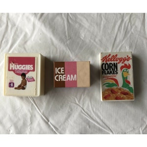 Op de afbeelding: Drie miniatuurdozen met productverpakkingsontwerpen. De eerste doos is wit met roze en blauwe accenten en zegt "Huggies UltraTrim" met een baby erop. De tweede doos is bruin en roze en zegt "ICE CREAM". De derde doos is rood, geel en groen en zegt "Kellogg's Corn Flakes The Original Best".
