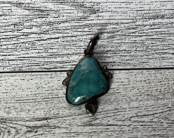 May include: A turquoise pendant with a silver-colored metal setting. The pendant has a teardrop shape and a dark, mottled pattern. The metal setting has decorative elements and a loop for a chain.