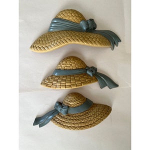 May include: Three wall decor pieces shaped like straw hats with blue ribbon accents. The hats are different sizes and are arranged in a vertical stack.