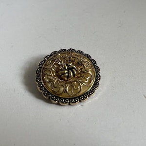 May include: A round, gold-toned brooch with a filigree design and a black center. The brooch has a scalloped edge.