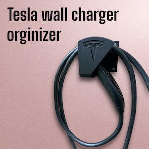 May include: Black Tesla wall charger organizer with a cable management system. The organizer has a Tesla logo and is mounted on a wall.
