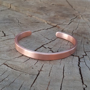 May include: A simple copper bracelet with a brushed finish. The bracelet is open and has a rounded shape.