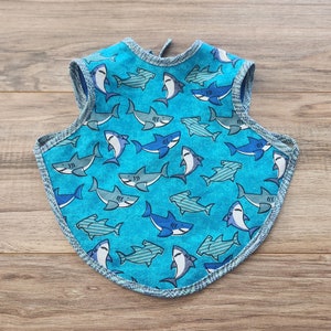 May include: A blue baby bib with a shark pattern. The bib has a tie closure and a scalloped edge.