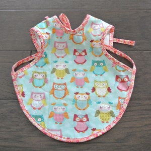 May include: A turquoise blue baby bib with a pink and orange trim. The bib is covered in a pattern of colorful owls.