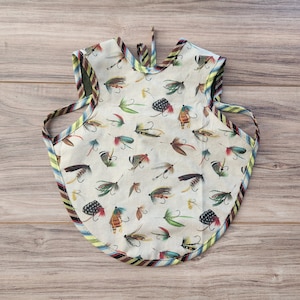 May include: A white bib with a colorful pattern of fishing flies. The bib has a green, brown, and blue trim.