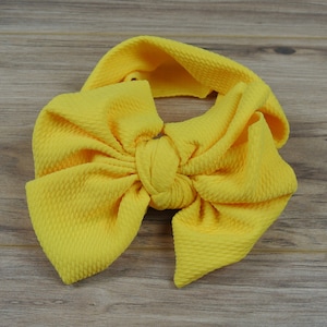 May include: A vibrant yellow baby headband featuring a large bow. The headband is crafted from a textured material and knotted at the centre. The bow is substantial and eye-catching, with a soft, rounded form. The headband rests on a wooden surface.