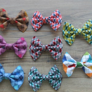 May include: Nine fabric bow ties in various patterns and colors are displayed on a wooden surface. The bow ties feature argyle and diamond patterns in shades of brown, pink, red, gray, blue, green, and multi-colored designs. The bows are neatly arranged.