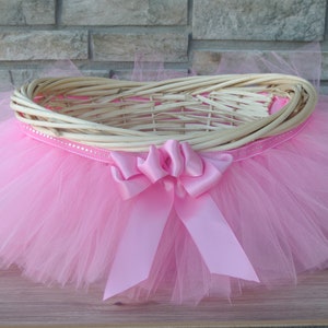 May include: A pink tulle tutu with a wicker basket in the center. The basket has a pink satin ribbon tied around it with a large bow in the front.