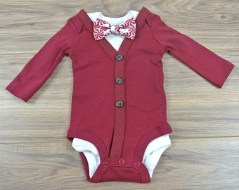 May include: A burgundy baby bodysuit with a white shirt collar and a red and white peppermint bow tie. The bodysuit has long sleeves and three brown buttons down the front. The outfit is designed to look like a suit.