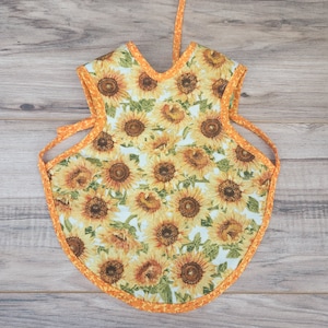 May include: A white bib with a sunflower print and orange trim. The bib has two ties that can be tied around the neck.