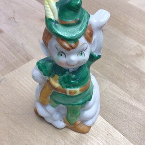 May include: A ceramic figurine of a whimsical elf wearing a green hat with a yellow feather, a green and brown outfit, and a white cloud-like base.