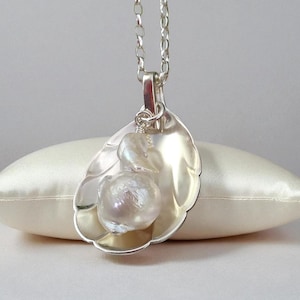 May include: A silver pendant necklace with a leaf-shaped design, holding a large, irregular pearl. The pendant is suspended from a fine silver chain. The pearl has a lustrous, off-white hue. The necklace is presented on a cream-coloured cushion.