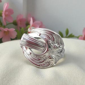 May include: A silver ring crafted from a spoon, featuring ornate floral and scrollwork details. The ring's design incorporates the spoon's handle and bowl, creating a unique and vintage-inspired accessory. The ring is displayed on a white surface.