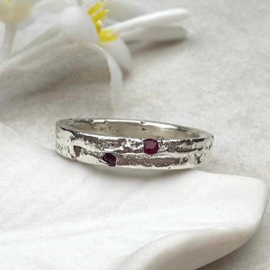 May include: A textured silver ring with two small, round, red gemstones. The ring is set against a white background with a white flower in the upper left corner. The ring's surface has a rough, organic texture.