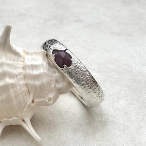 May include: A textured silver ring with a dark red gemstone. The ring is resting on a white seashell against a white background. The gemstone is a deep, rich colour, and the silver has a hammered appearance.