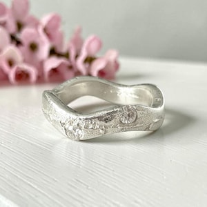 May include: A silver ring with a wavy, organic band featuring a textured, matte finish and two small, clear, round-cut stones embedded into the metal surface. The ring has an irregular, undulating shape that follows a gentle, flowing curve.