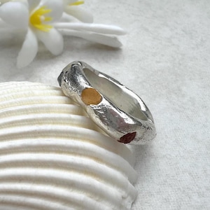 May include: A silver ring with a textured surface and a unique design. The ring features a small, round, gold-colored accent and a small, red-colored accent. The ring is displayed on a white seashell, with a white flower in the background.